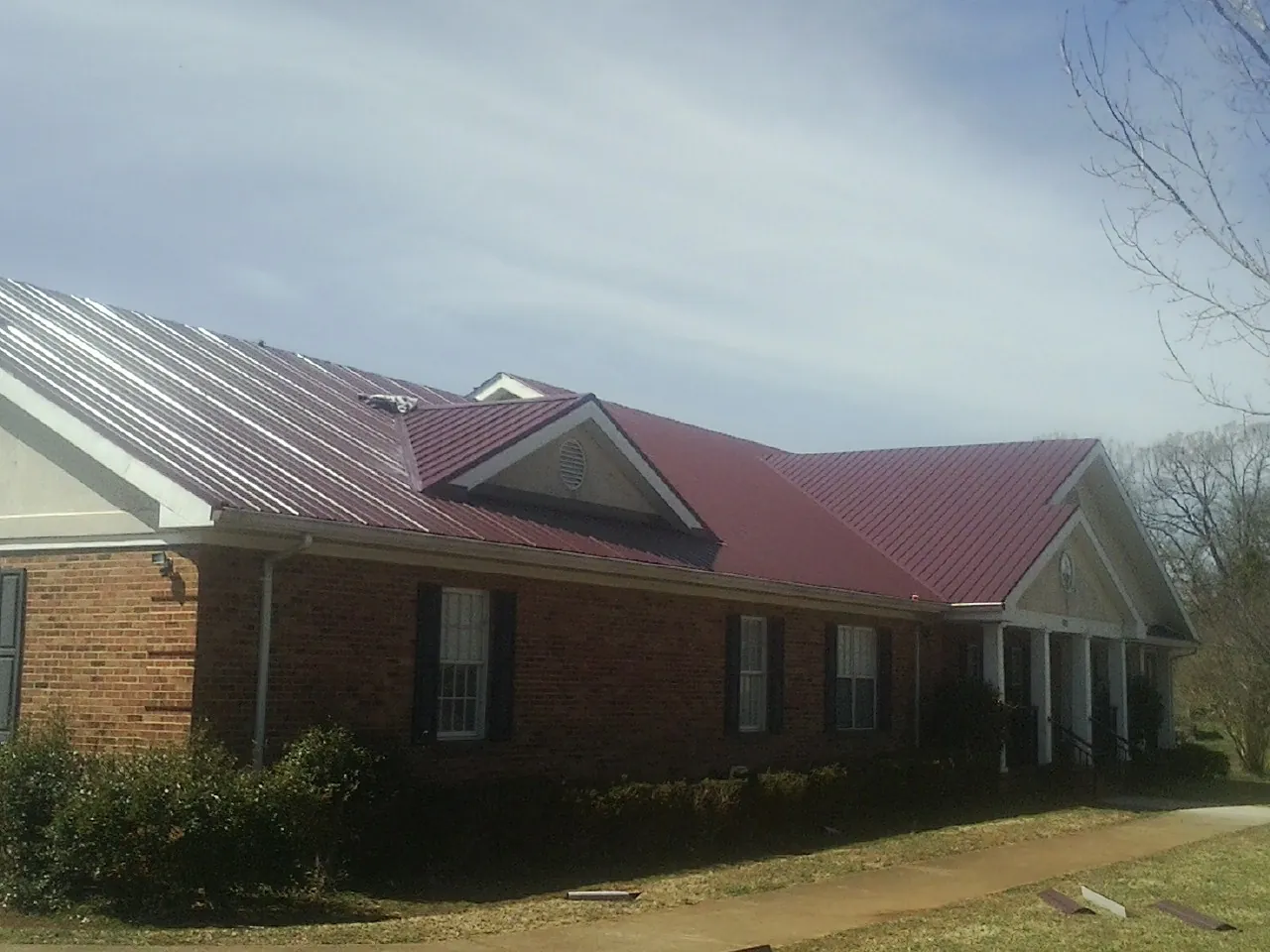Red metal roof with gabled entries on a long brick building, from a Hail Damage Roof Repair job in Oyster Bay
