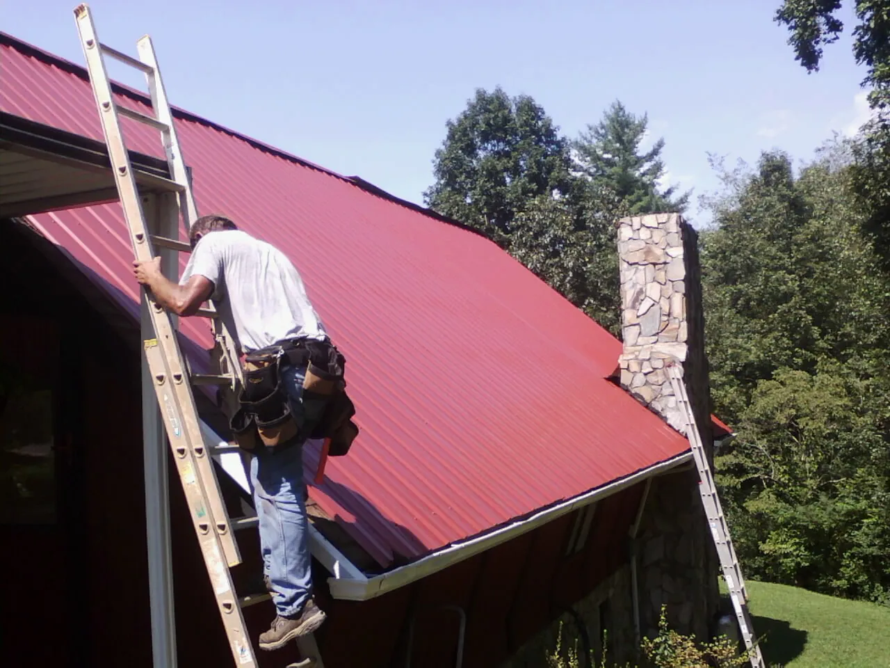 Roofer on a ladder beside a red metal roof and stone chimney — Soffit & Fascia Repair by Roofing Brothers in Oyster Bay